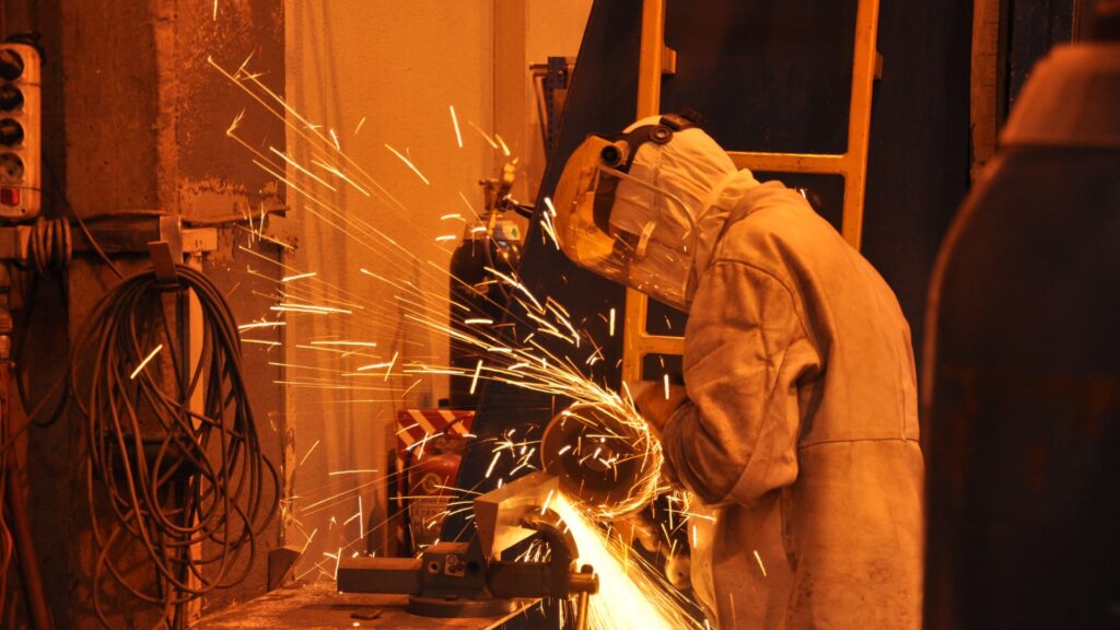 A person wearing a protective welding helmet and jacket is working with metal, creating a shower of bright orange sparks in a dimly lit workshop.